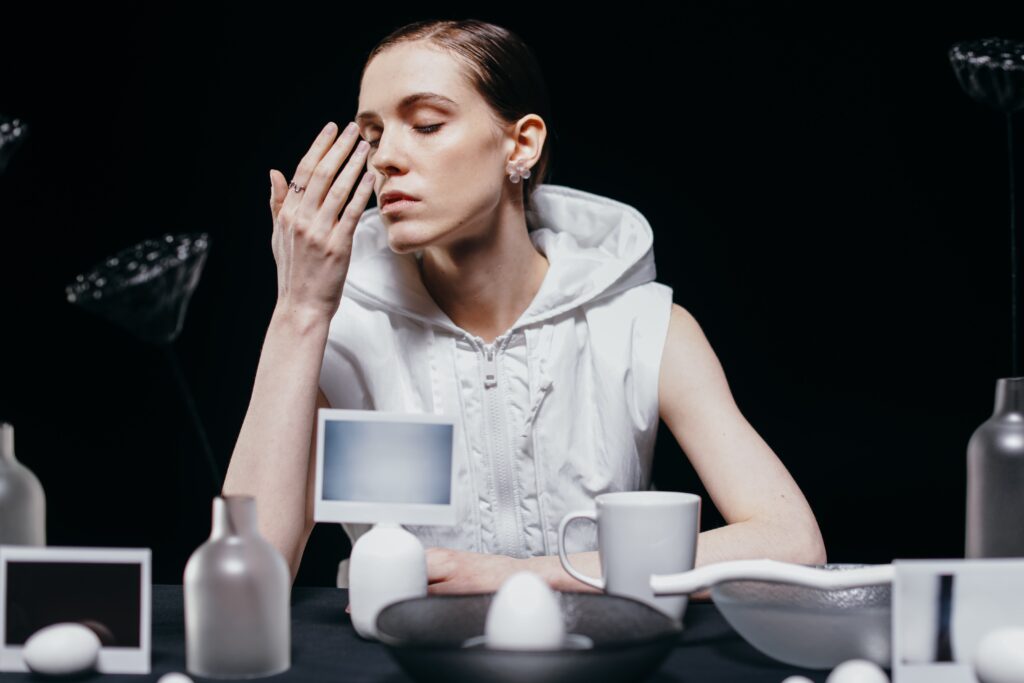 Woman in white vest smelling her hand with her eyes are closed. Various objects, such as a mug, a polaroid, an egg, are laid across a table in front of the woman.