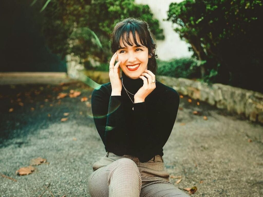 A woman sits outside on pavement. She is smiling and looking off from the camera with her hands touching her face.