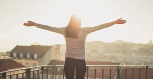 A woman stands on her balcony outlooking the rows of townhouses. She throws her arms wide open with the setting sun framing her silhouette