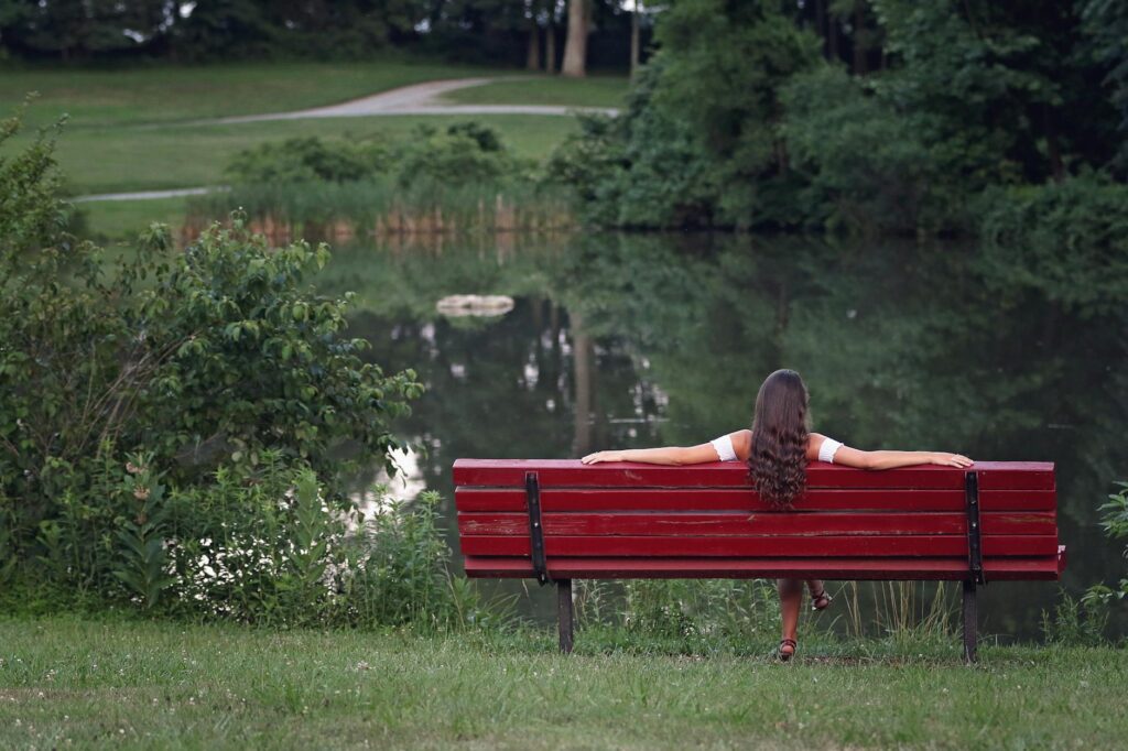 A woman sits relaxed with arms splayed out on a red bench with the back to the camera. The bench overlooks a pond and green brush.