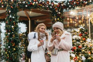 Two women in fuzzy white coats enjoy a holiday cup of beverage in front of a holiday decorated frame.