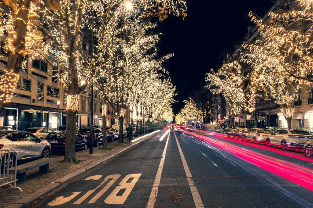 A long exposure of cars along a road in the night. The trees lining each side of the road are lit up with white holiday lights.