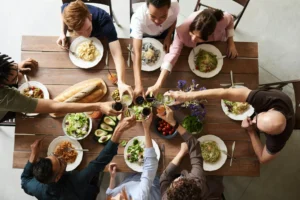 Several people sit around a wooden table with plates full of food. The photo is take from above, capturing their arms reaching into the middle and clinking their drinks together.