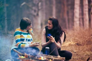 Two woman sit around a campfire with mugs, smiling and talking to one another.