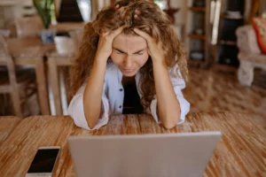 A woman sits at a table with her heads in her head and brows pinched. Her laptop and phone sit in front of her. She's overwhelmed.