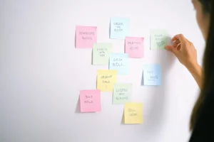 A person is placing a sticky note to the wall filled with other colored sticky notes. Each note carries a different reminder: "buy milk," "urgent call," Listen to new albums," "call mom."