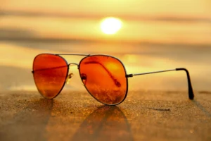 A pair of red tinted sunglasses rest on the sand. The sun sets behind it.