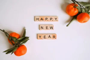 "Happy New Year" is spelled out in scrabble letters on a white background. A couple pieces of fruit frame it.