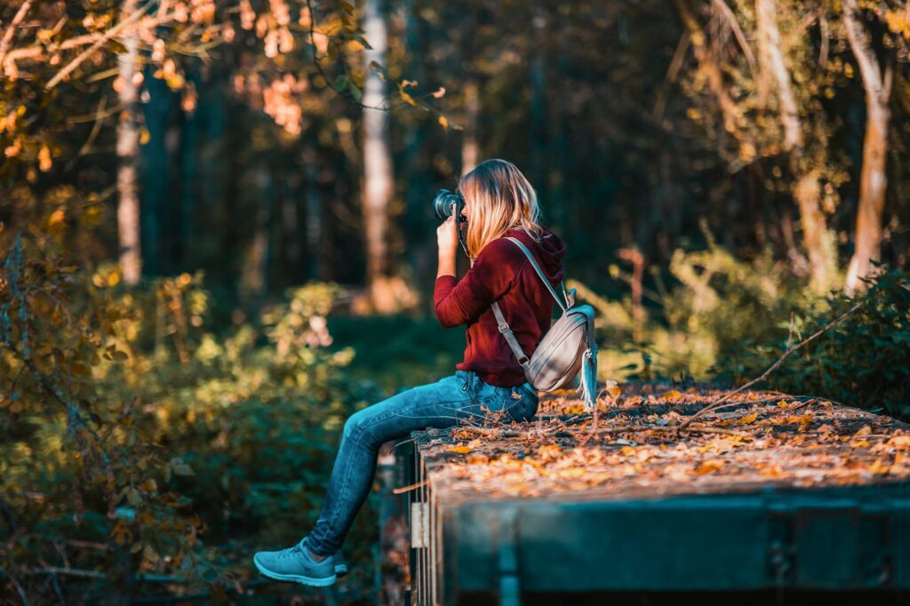 A woman in a red long-sleeve and jeans sits on the edge. She faces away from the camera with her own camera held up to her face. Woodsy greens and trees fill in the background.