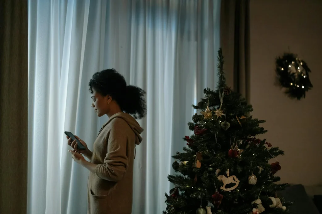 A woman stands with her back to a decorated christmas tree. She clutches a phone in her hand, looking at it in sorrow.