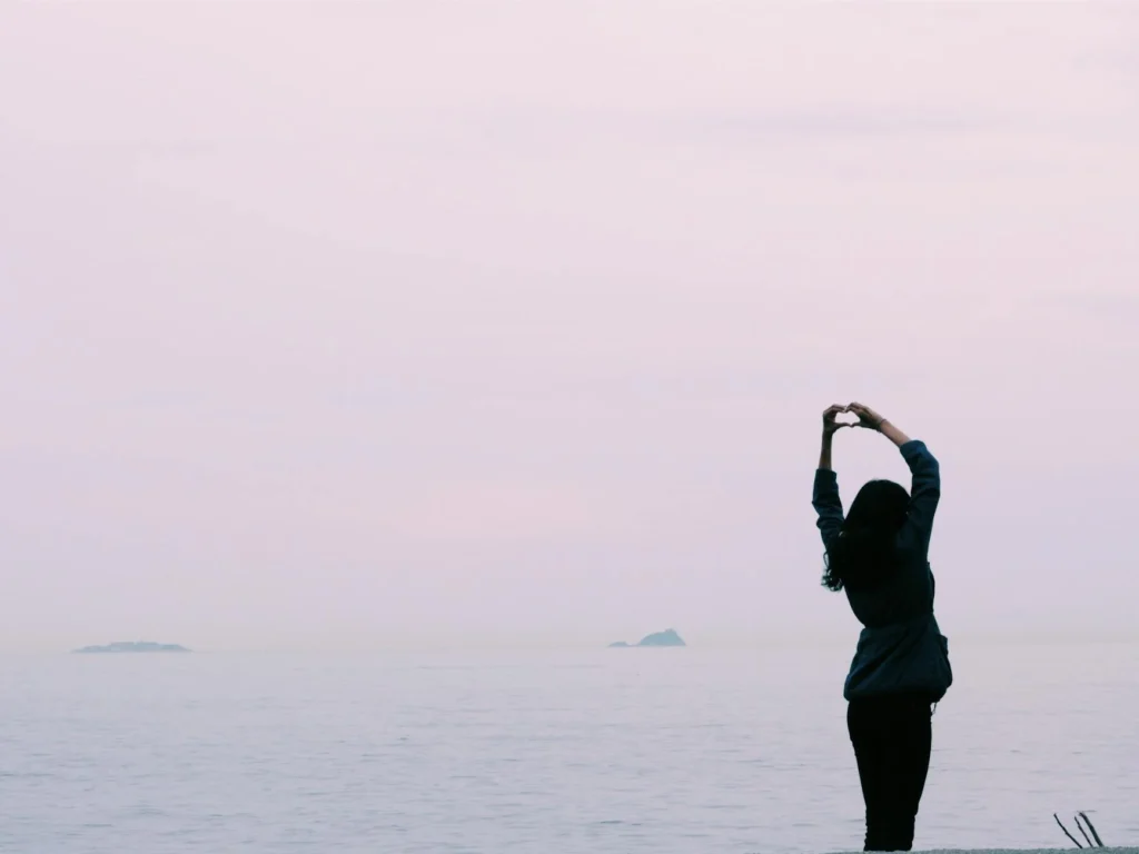 Against a pale sea and sky background, a woman stands on the shore. Her hands reach up and form a heart.