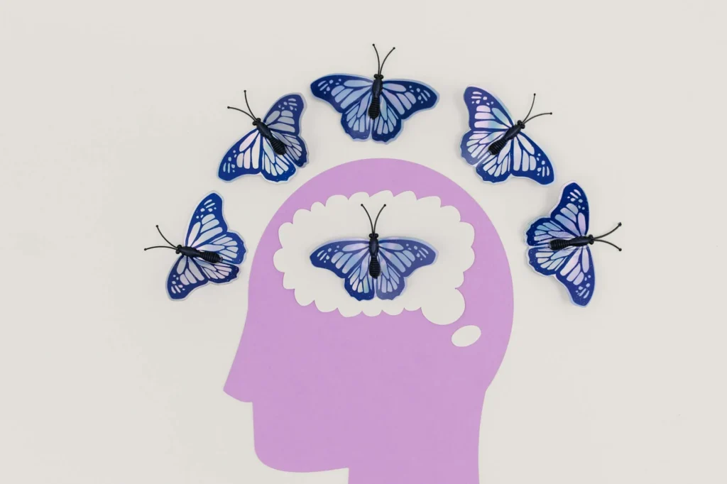 A pink silhouette cut out of a head. A white cut out of a thinking bubble is set over the head and contains a butterfly. Surrounding the head are 5 similar butterflies.