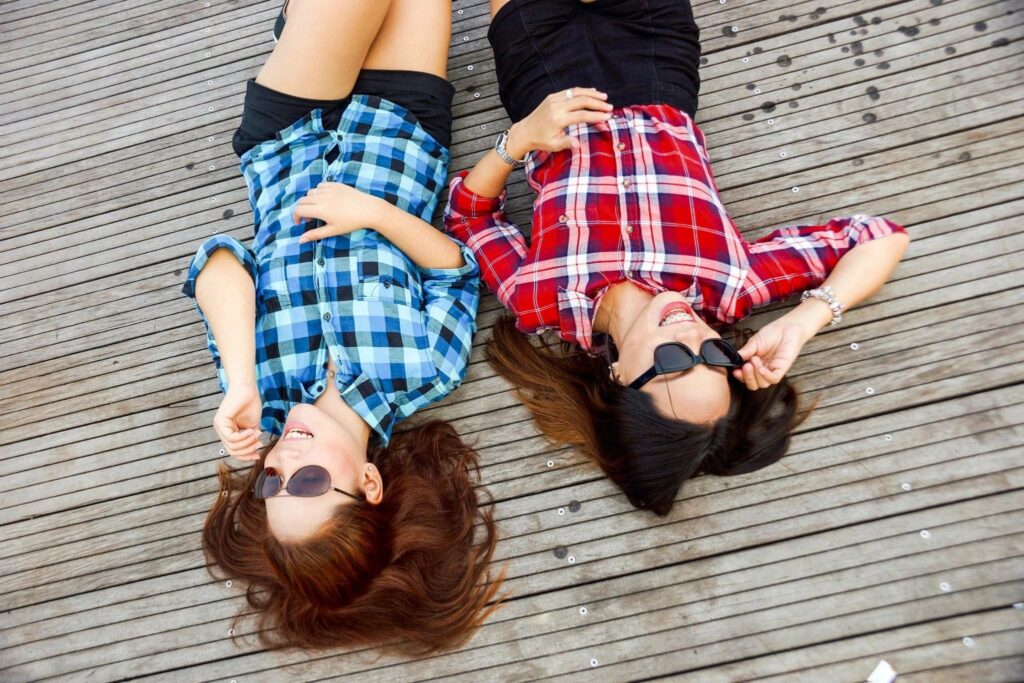 Two girls in blue and red plaid shirts lie on a deck side by side. They appear to be laughing.