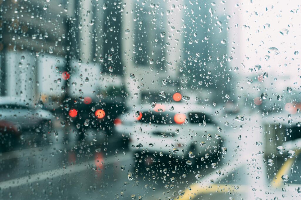 Rain drops cover the image as though looking through a window with rain dripping down. It faces into a street of blurry cars.