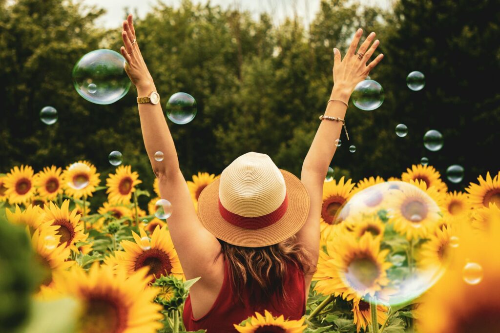 A woman throws her hands into the air as she's standing in a tall sunflower field. Trees frame her and bubbles are floating around the field.
