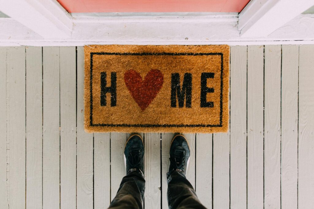 A topdown view of a person's feet standing in from of a welcome met that says home, where the "o" in home is a heart.