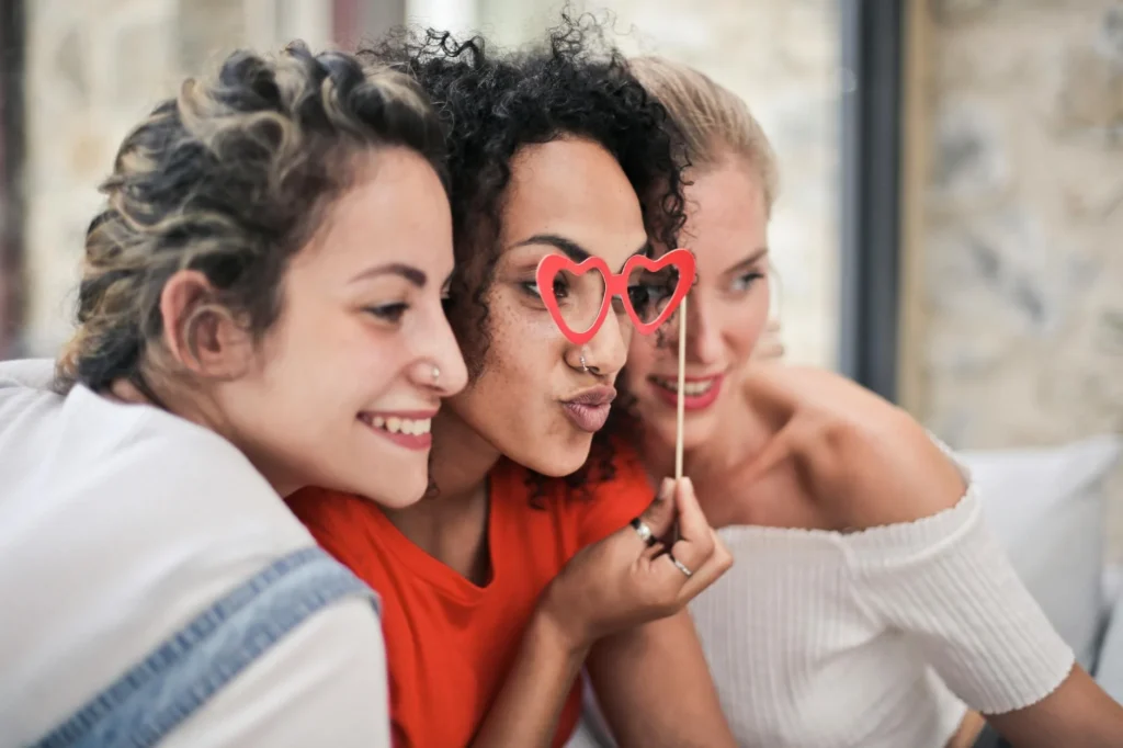 Three woman huddle together and look off to the side to an off-screen photographer. The middle woman holds up a pair of heart glasses.