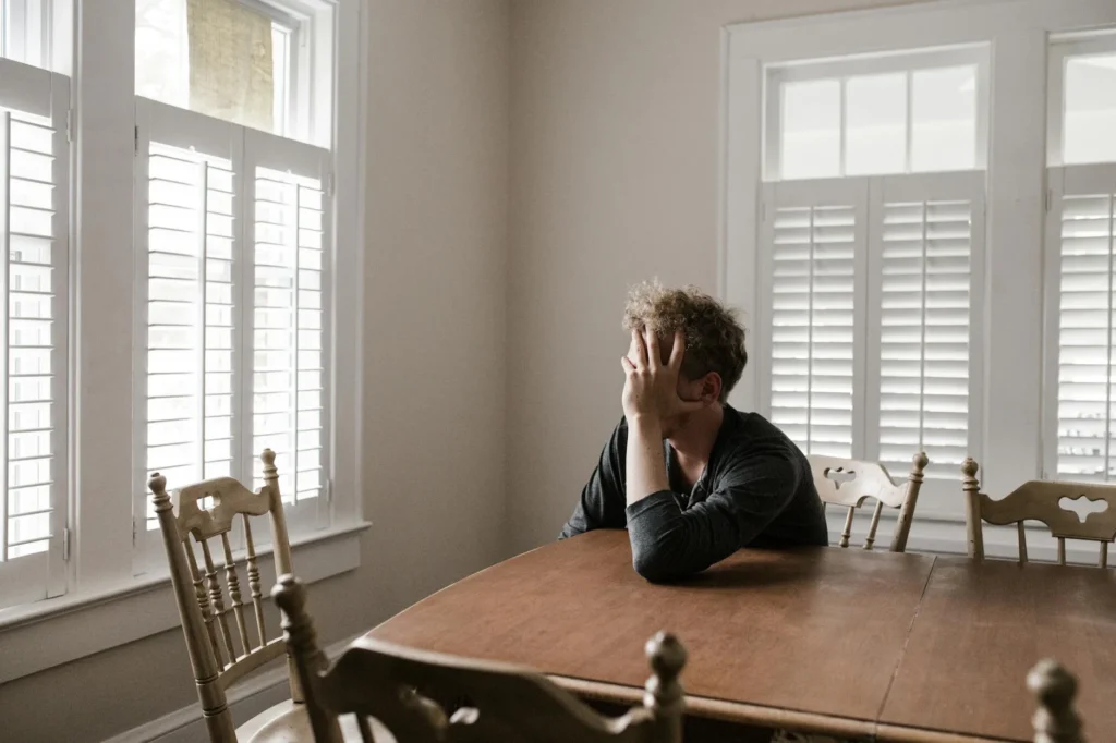 A man sits at a dining table, slightly slumped with his hand covering his face, looking defeated.